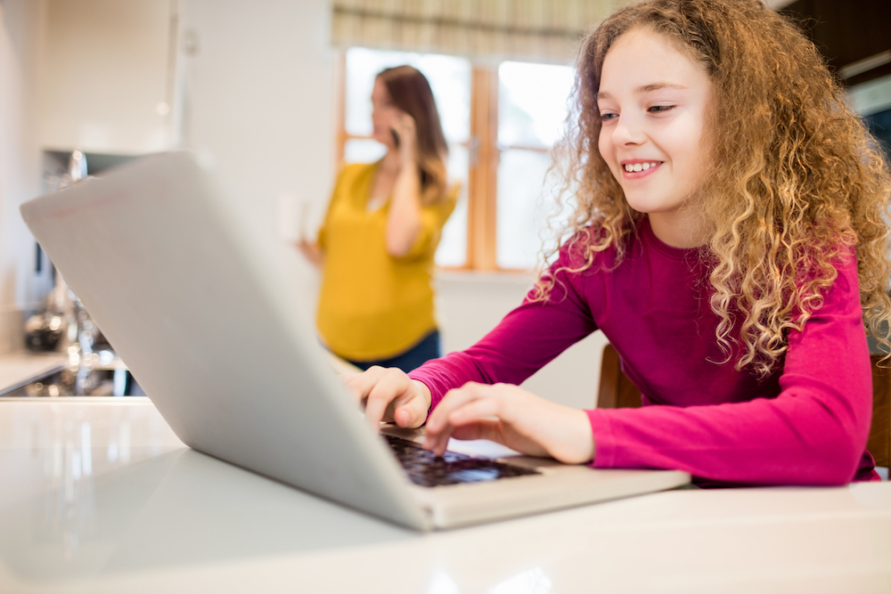 dziewczynka pisząca na klawiaturze komputera, a w tle stojąca kobieta rozmawiająca przez telefon